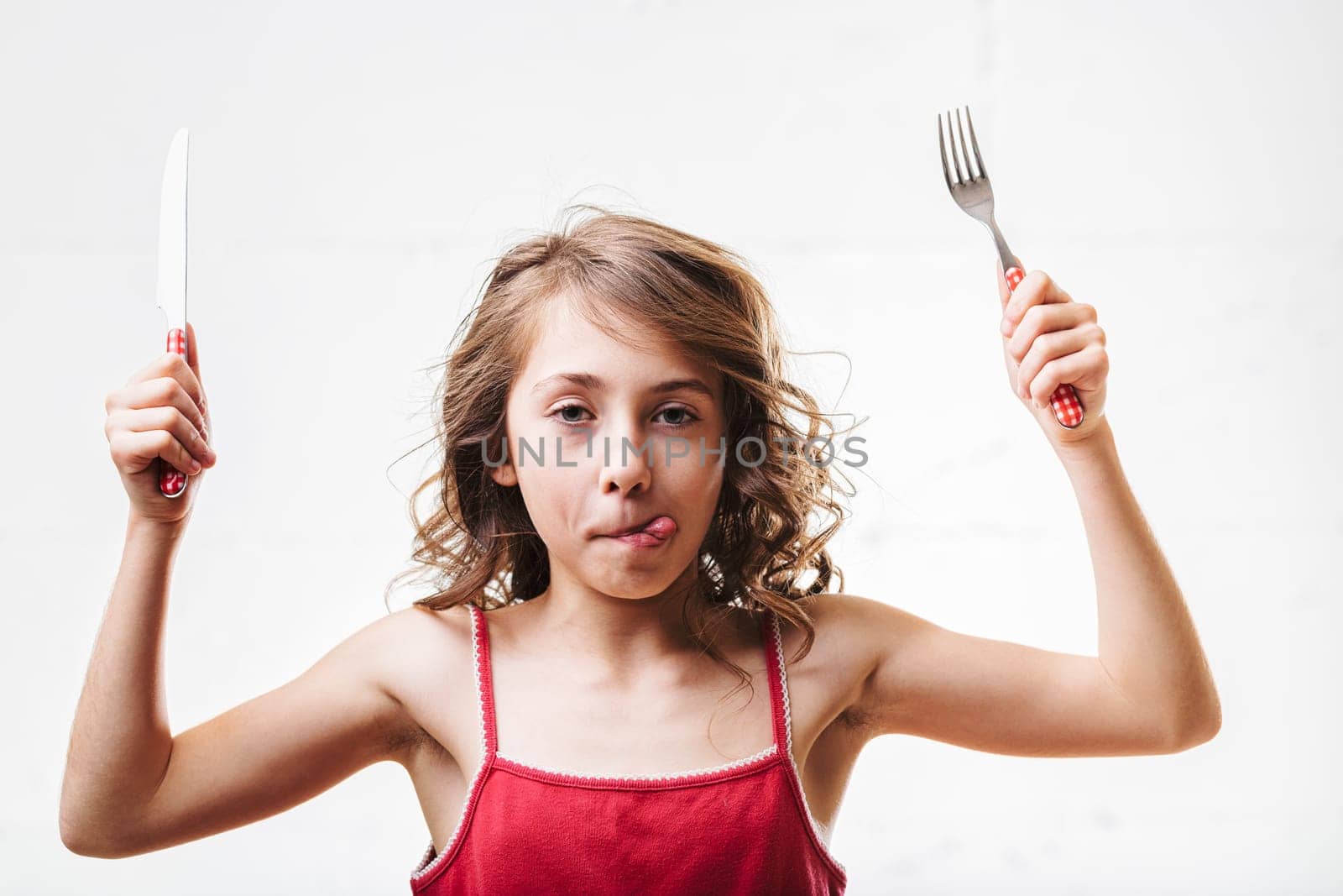 Young girl energetically holding a fork and knife, playfully sticking out her tongue, radiates excitement and joy in a lively mealtime setting