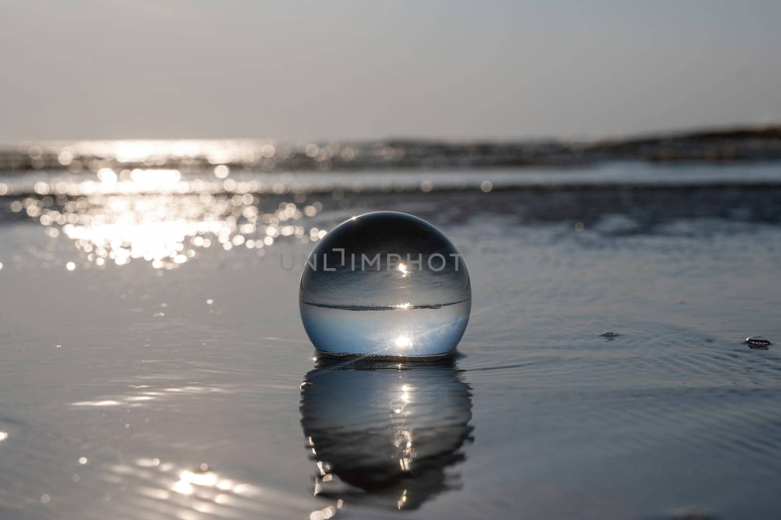 A glass ball lies in the waves on the sandy beach, the sea and the setting sun are reflected in the ball