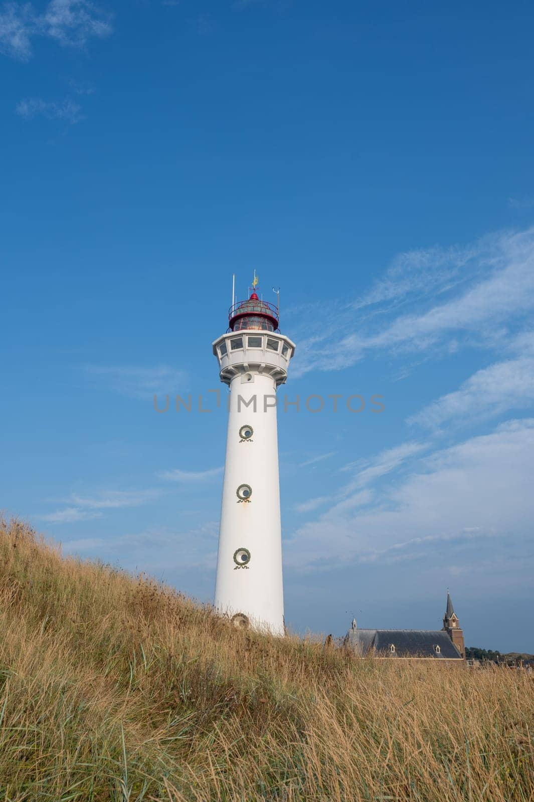 The approx. 28 m high white lighthouse J.C.J. van Speijk in Egmond aan Zee, North Holland, Netherlands with the Catholic St. Agnes Church, beach grass and sand dunes