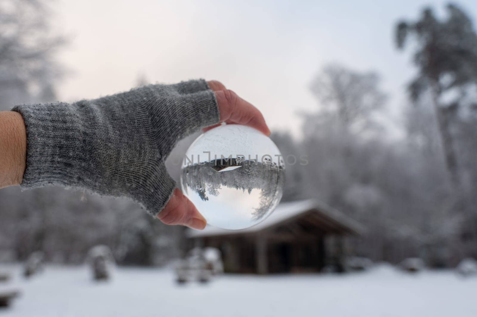 Human hand with a grey glove holds a glass ball reflecting a snow-covered landscape and a hut