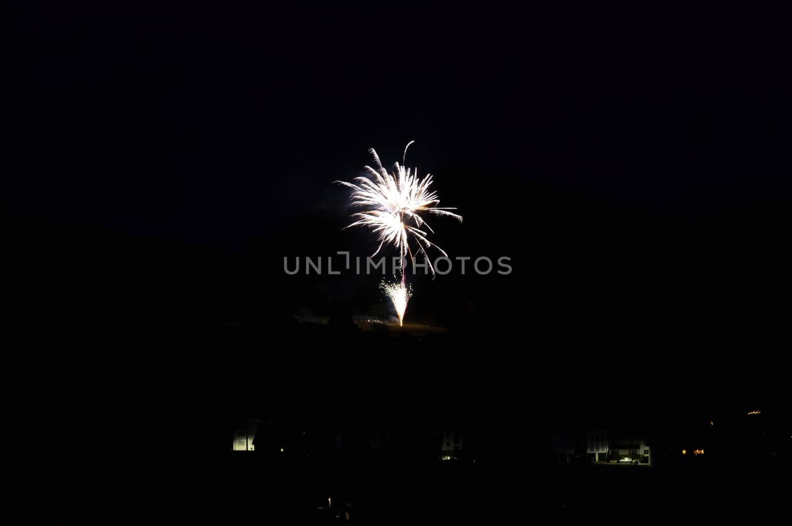 A firework rocket in the sky on a mountain at night, a few illuminated houses in the foreground