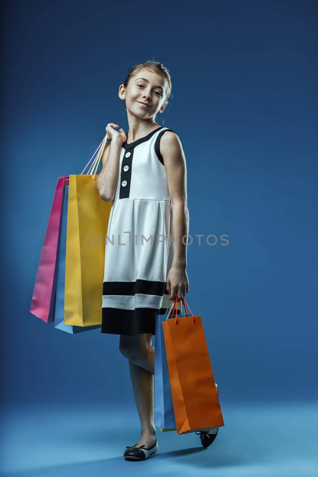 Young girl joyfully carrying colorful shopping bags, celebrating a successful shopping spree against a vibrant blue backdrop, radiating happiness and excitement