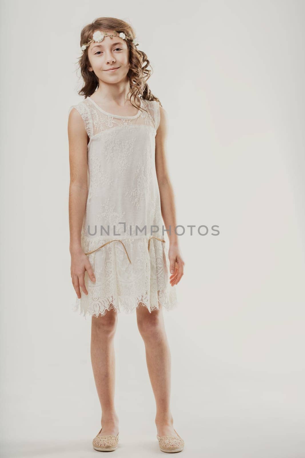 Smiling preteen girl wearing a delicate white lace dress and a charming flower crown, looking directly at the camera with a joyful expression in a studio setting