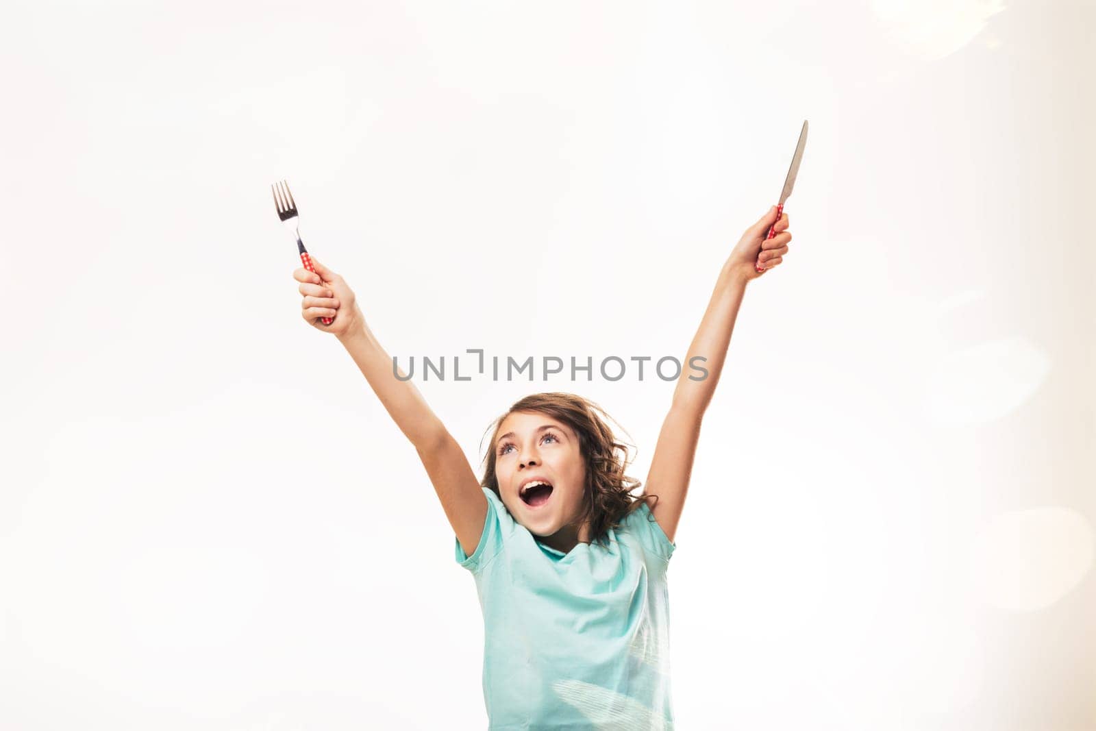 Enthusiastic preteen girl raising her arms, holding a fork and knife, expressing excitement and anticipation for a meal, against a bright white background