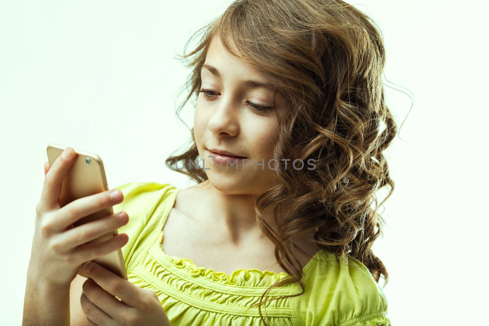 Portrait of a smiling preteen girl with curly, brown hair, holding a smartphone and engaging with the screen, set against a clean white background