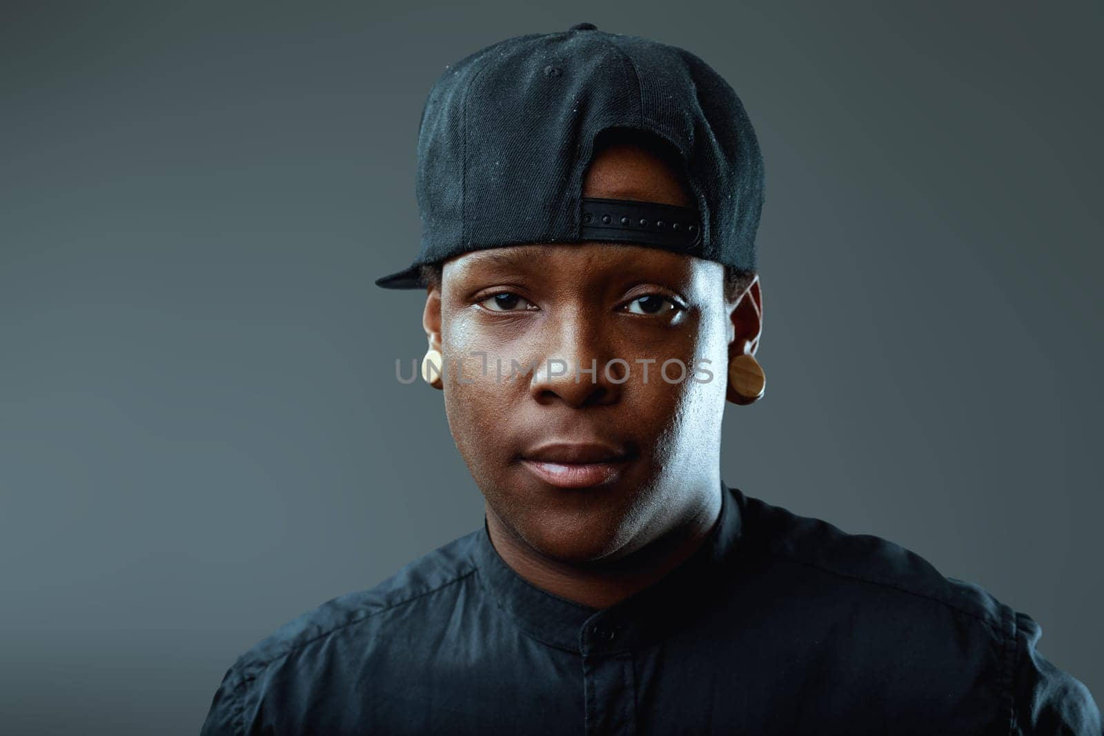Portrait featuring a serious young african-american man with ear plugs and a baseball cap, staring intensely at the camera against a moody gray background, exuding confidence and style