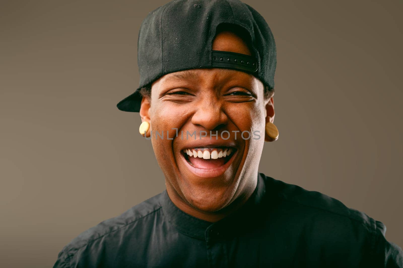 Cheerful young musician wearing a stylish black outfit and baseball cap, laughing joyfully and displaying a bright smile, radiating happiness and positive energy in a studio setting