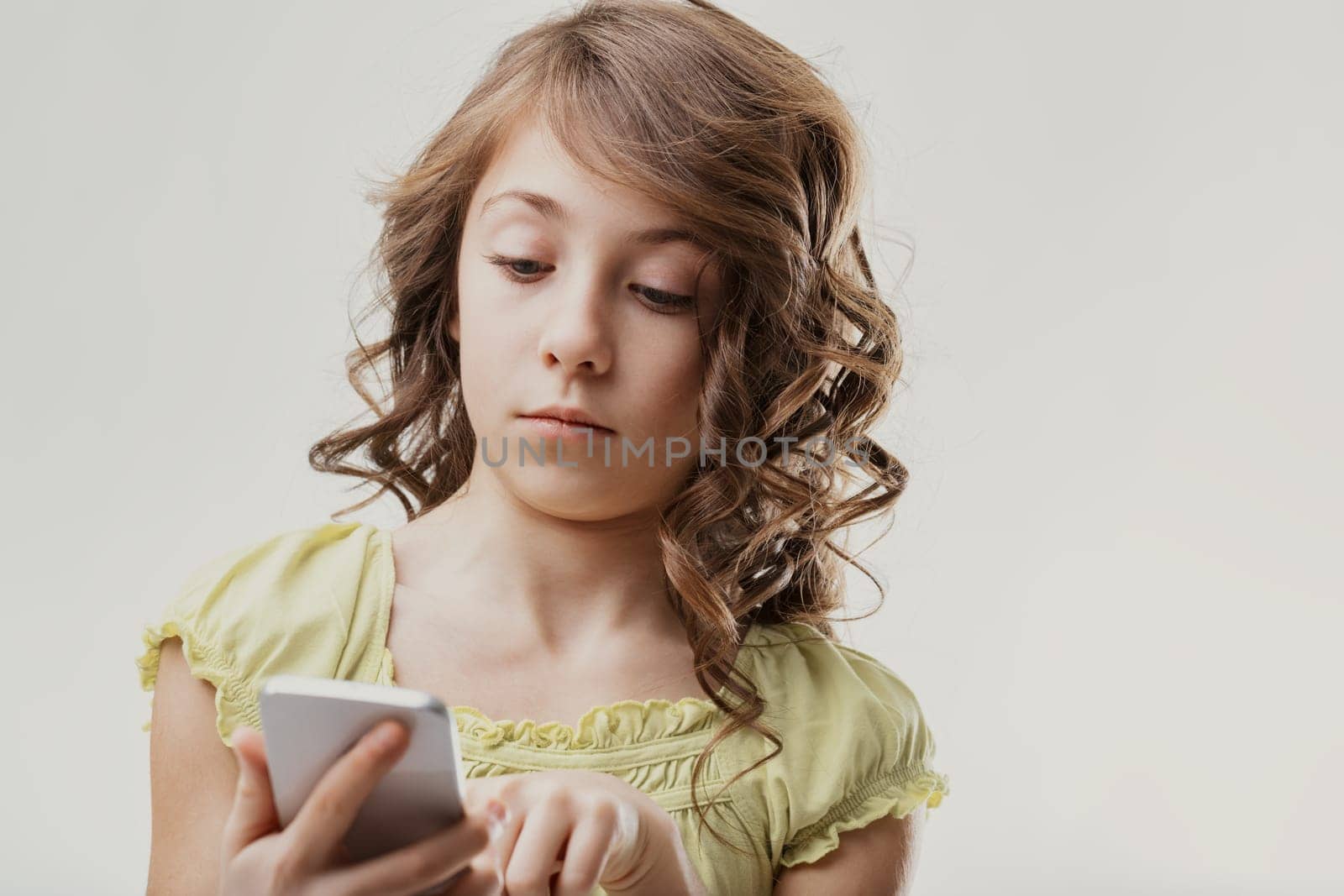 Elementary age girl with curly hair holding a smartphone and looking at the screen with a focused, pensive expression, suggesting concentration or contemplation