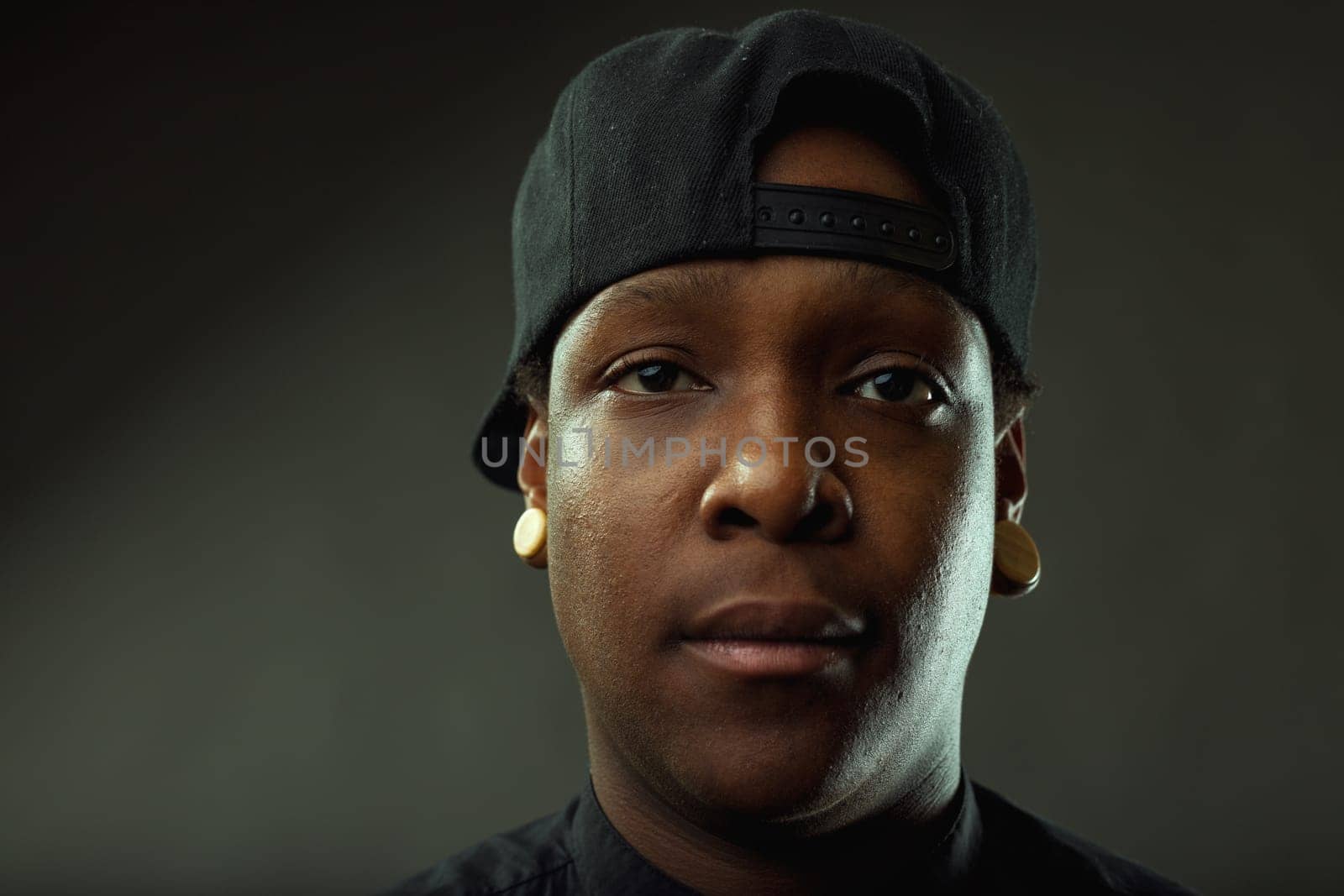 Close-up portrait of a serious young african american man wearing a baseball cap and plugs. Staring intensely at the camera against a dark background. Conveying a sense of confidence and urban style