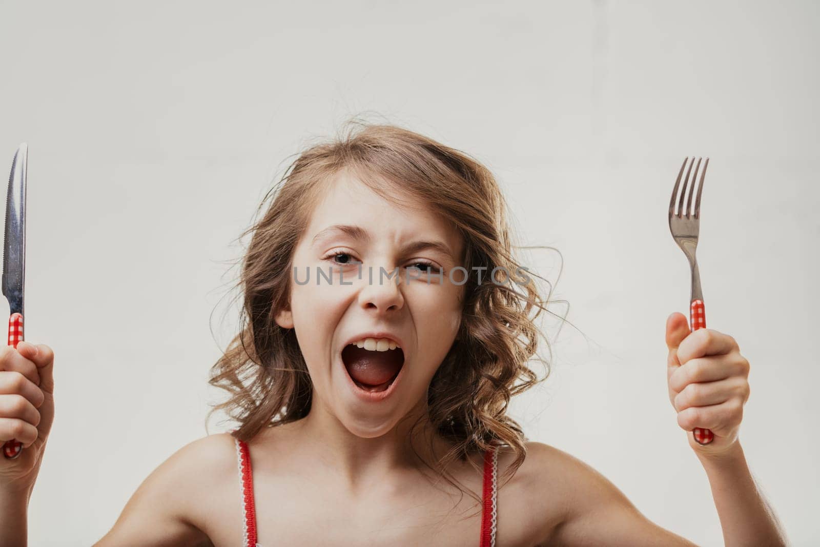 Young caucasian girl with brown curly hair holding a fork and knife, shouting with excitement against a white background, clearly expressing hunger and eagerness to eat