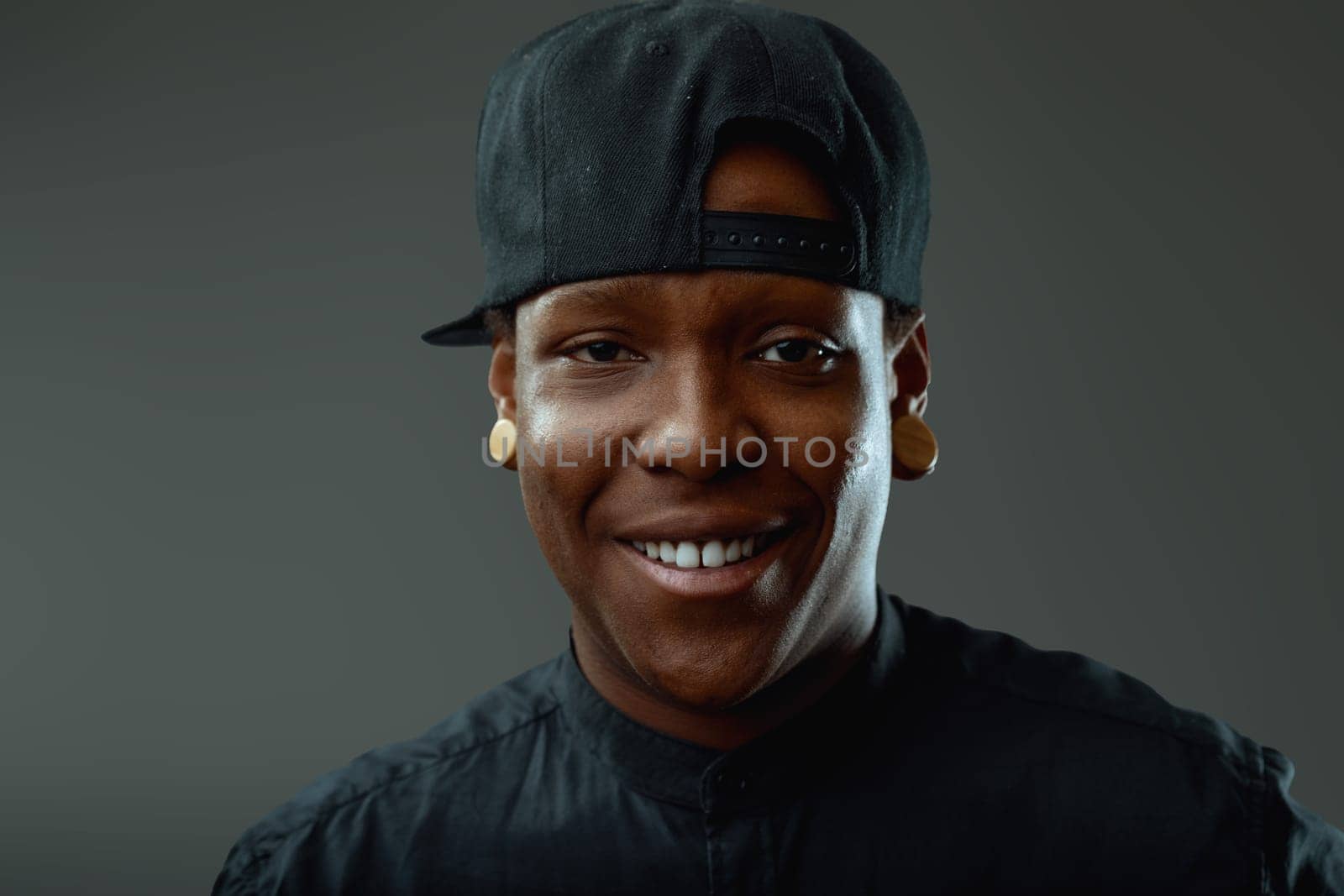 Studio portrait of a cheerful young african american man wearing a baseball cap and black shirt, smiling confidently at the camera against a gray background