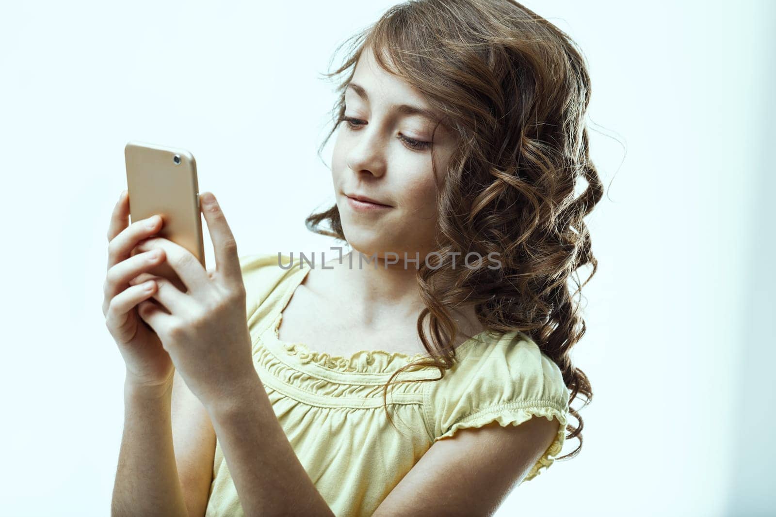 Cute elementary age girl with curly hair, smiling while holding and using a smartphone, engaged with the screen. Bright expression radiates happiness in a studio setting