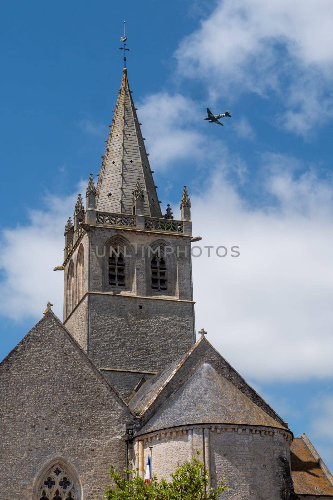 A church steeple with a plane flying in the sky. The plane is flying low and is in the background. Saint Come du Mont, normandy, France