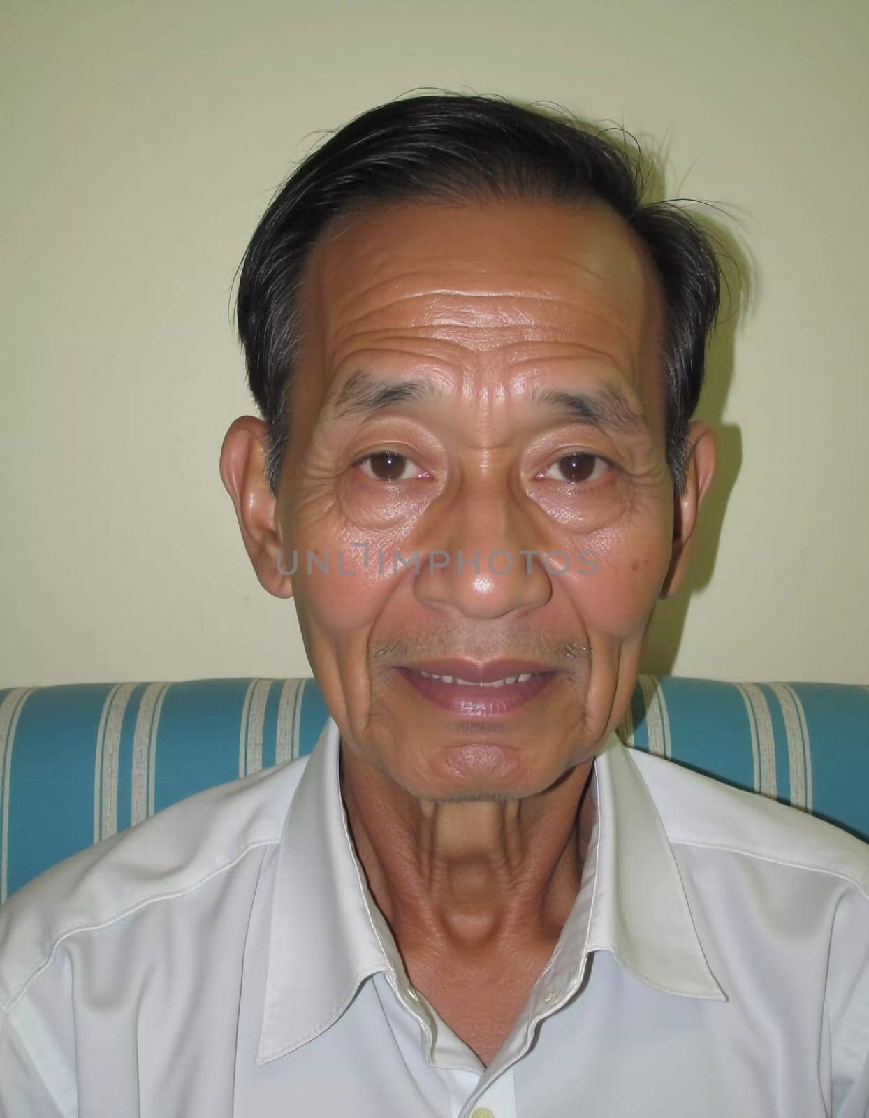 An elderly man residing in Thailand sits comfortably indoors, displaying a warm smile. His features reflect a life rich in experiences, showcasing wisdom and tranquility.