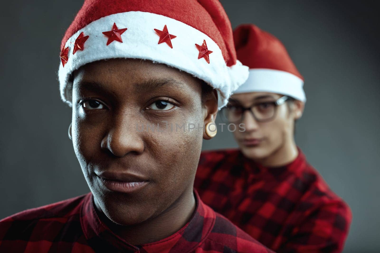 Two diverse young adults wearing matching red and black checkered shirts and santa claus hats looking at the camera with a serious expression