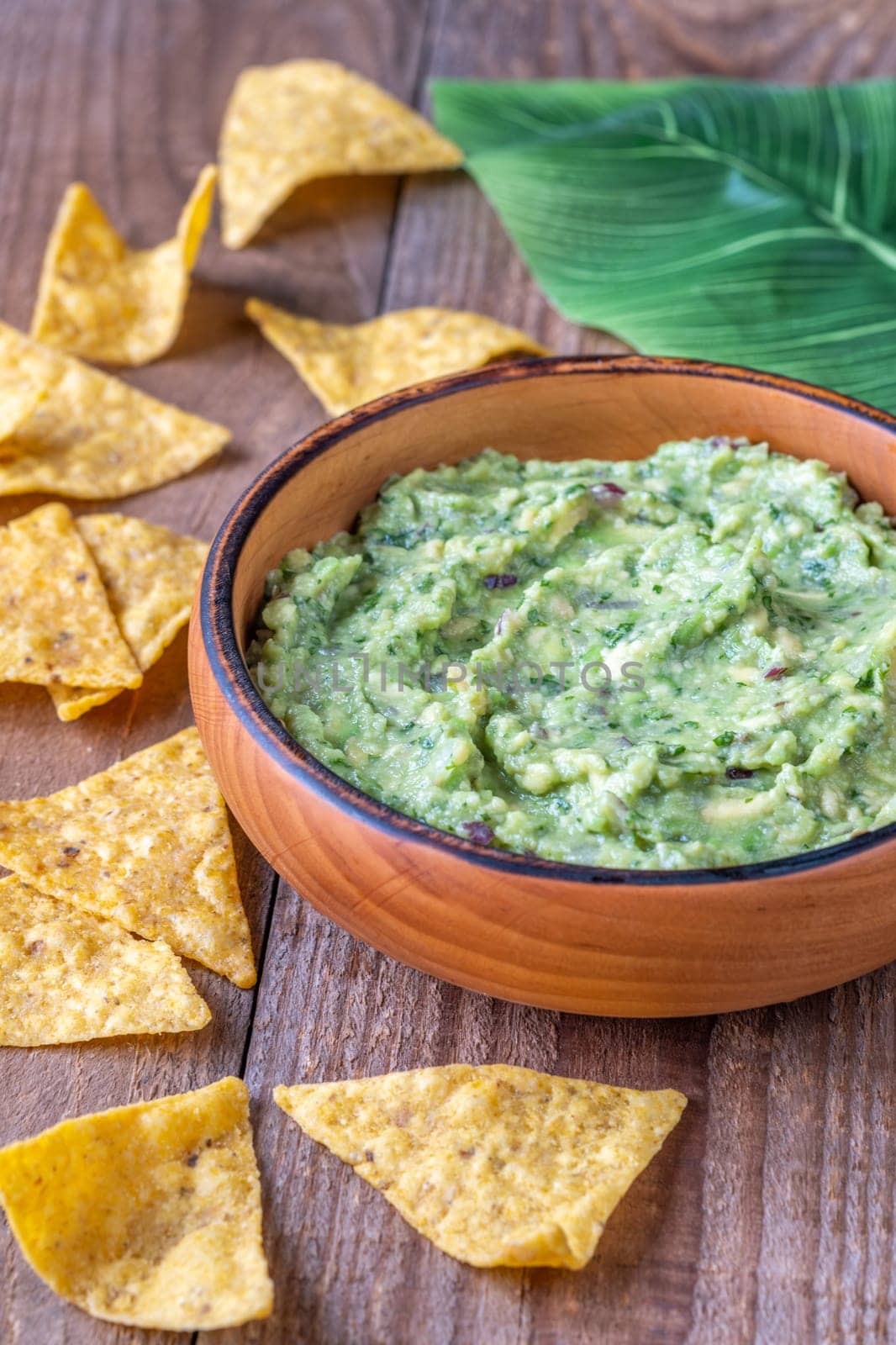 Wooden bowl of guacamole with tortilla chips and a large green leaf on a rustic wooden table
