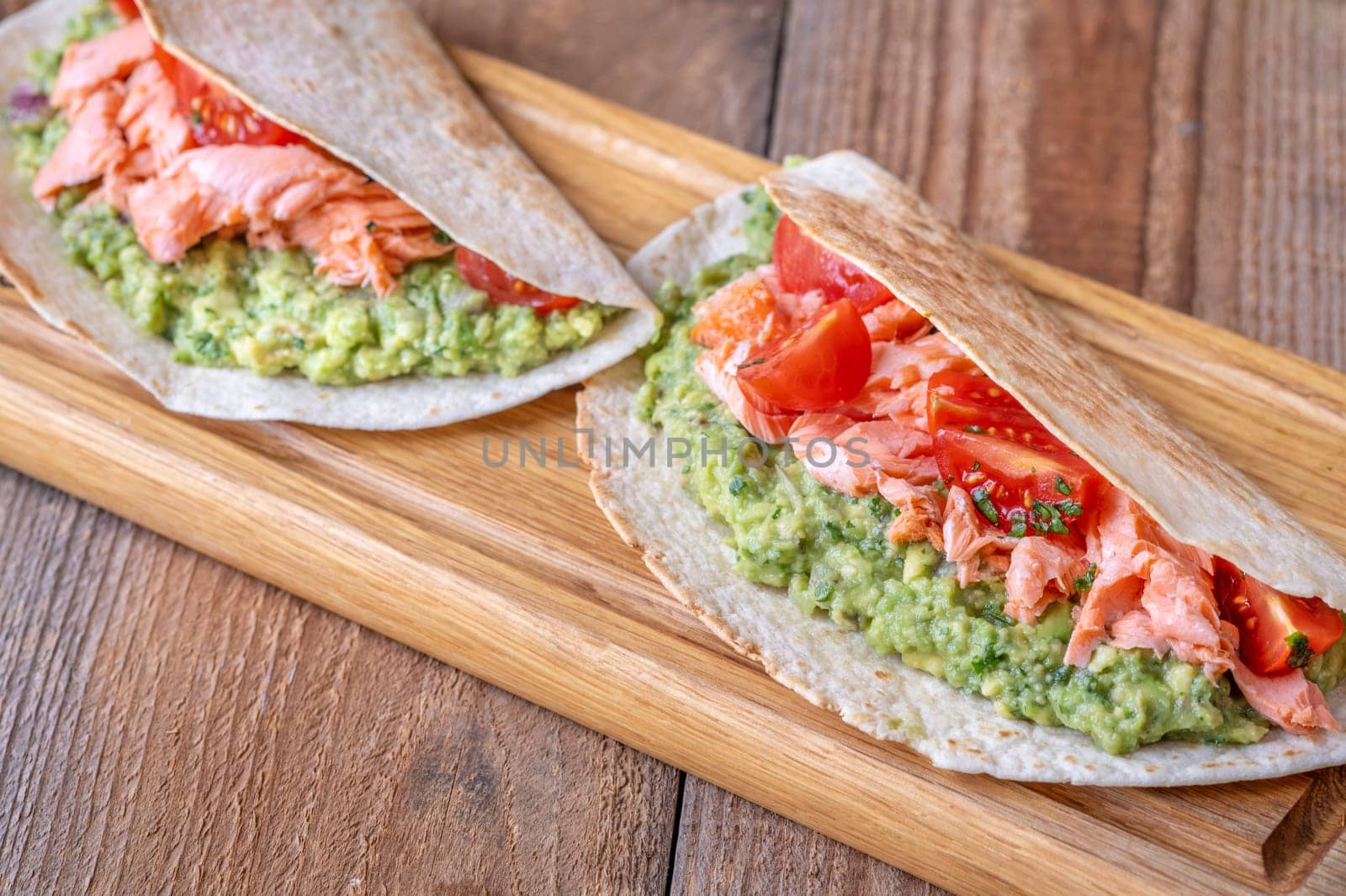 Two tacos filled with guacamole, baked salmon, and tomatoes on a rustic wooden table