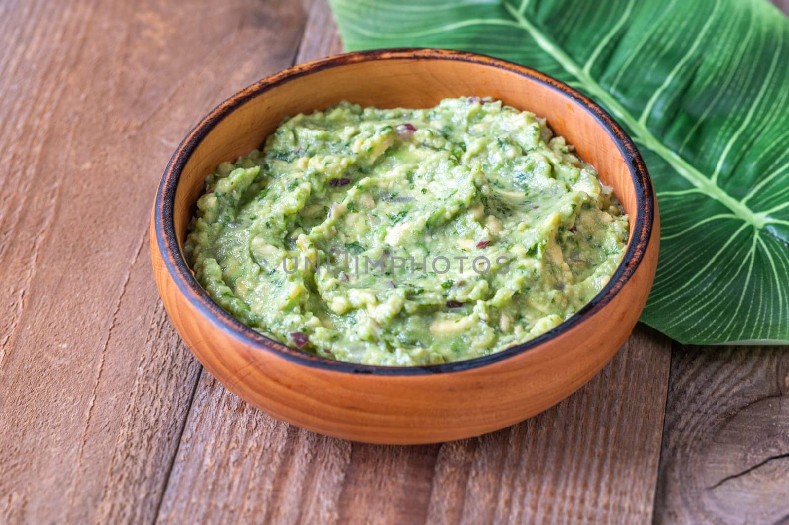 Wooden bowl of creamy guacamole on a rustic wooden table with a large green leaf