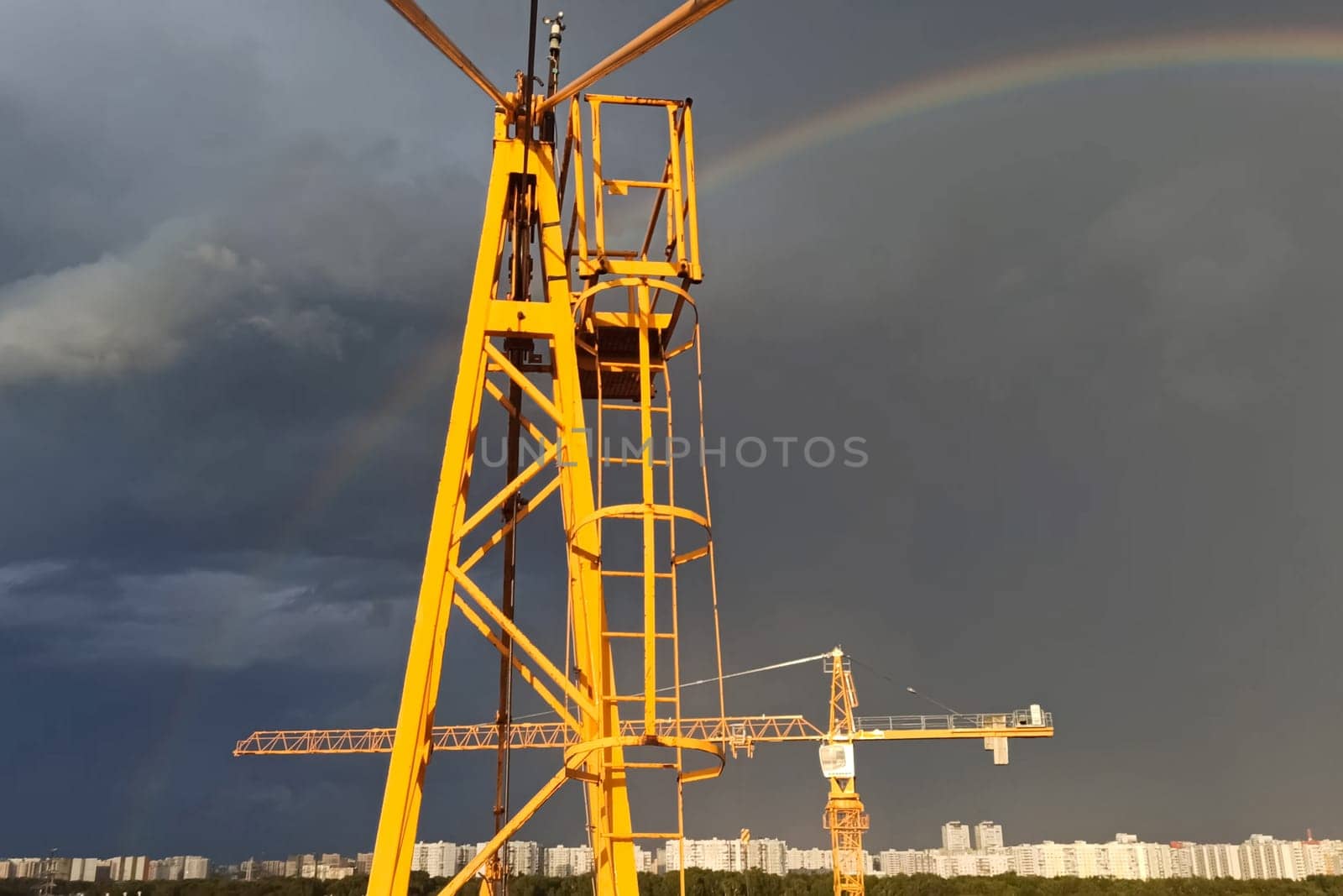 Tower cranes against the sky with clouds and rainbow.