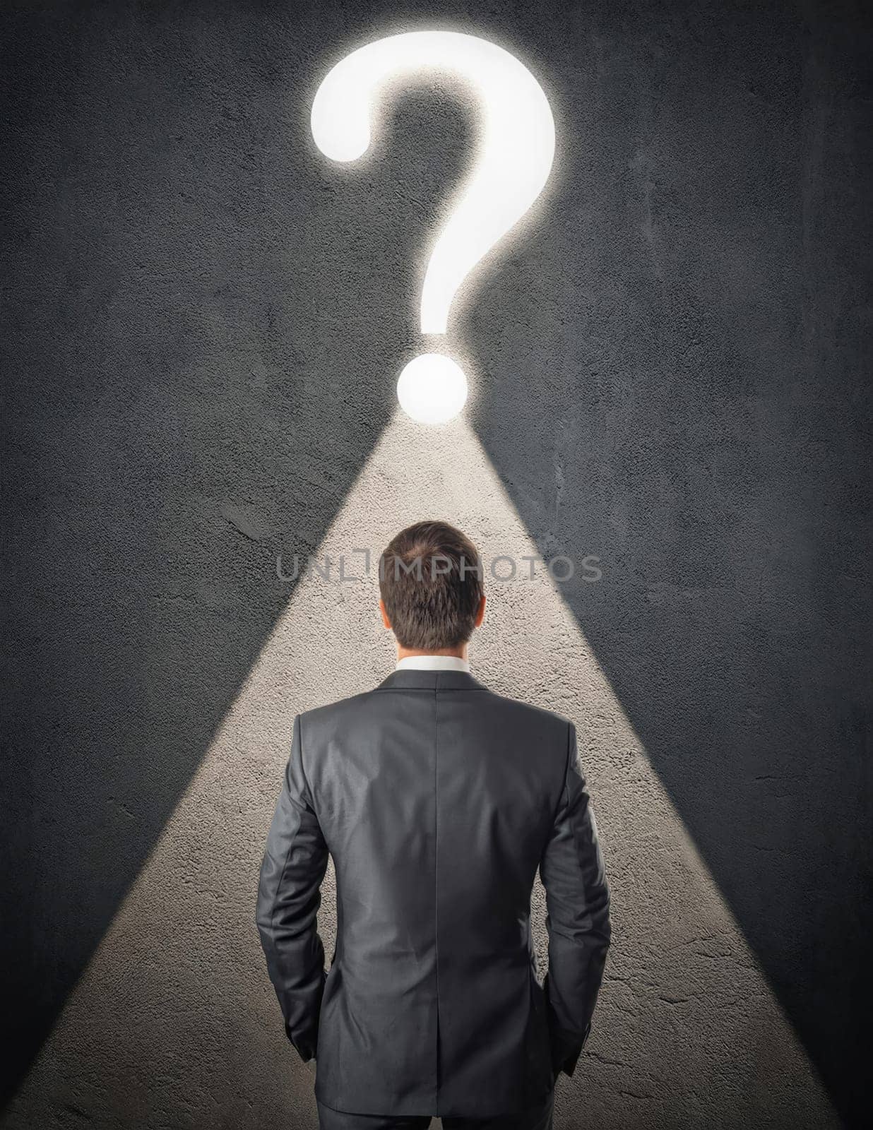 Rear view doubtful businessman inside a dark room, stands in front of a concrete wall with illuminated interrogation mark. Seeking answer to his questions. Contemplation concept