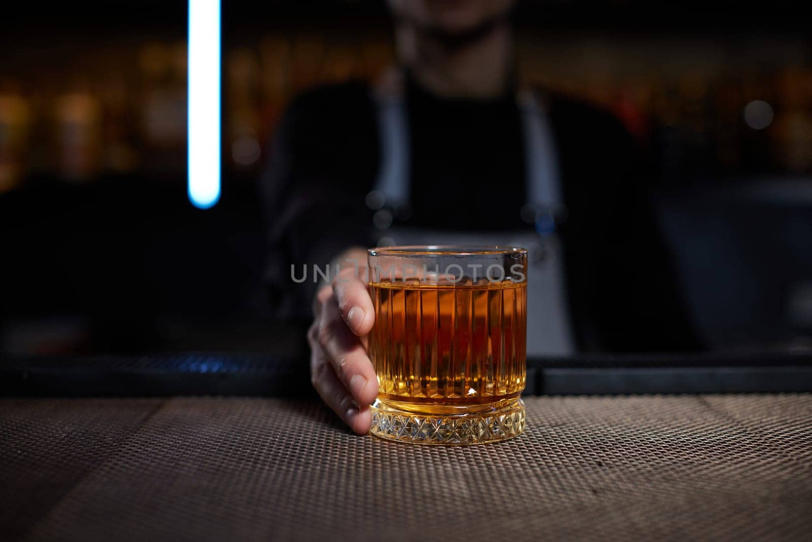 Barman holding out glass with alcoholic cocktail to the camera forward. Bartender is making cocktail