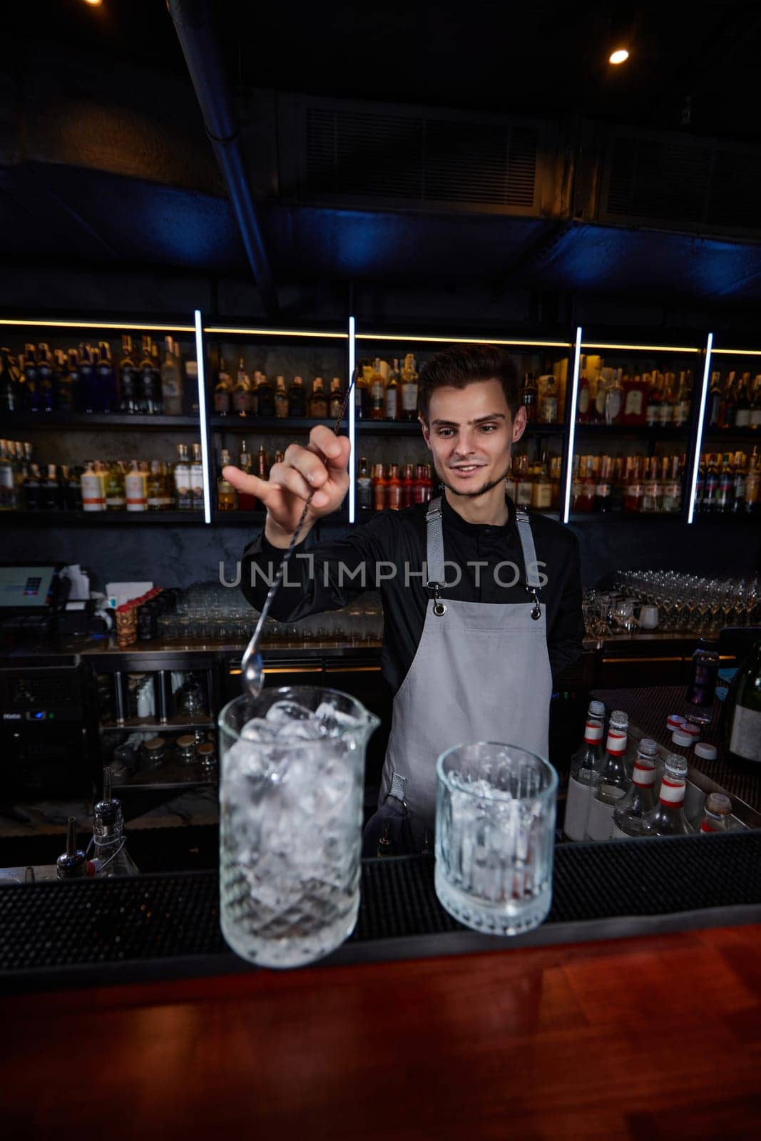 Barman in apron stirring ice cubes in cocktail glass with spoon at the bar