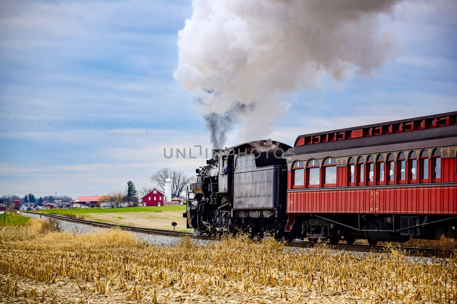 A steam train chugs along a country track amidst golden fields, releasing billowing smoke into the blue sky. Nearby, a classic red passenger car follows closely.