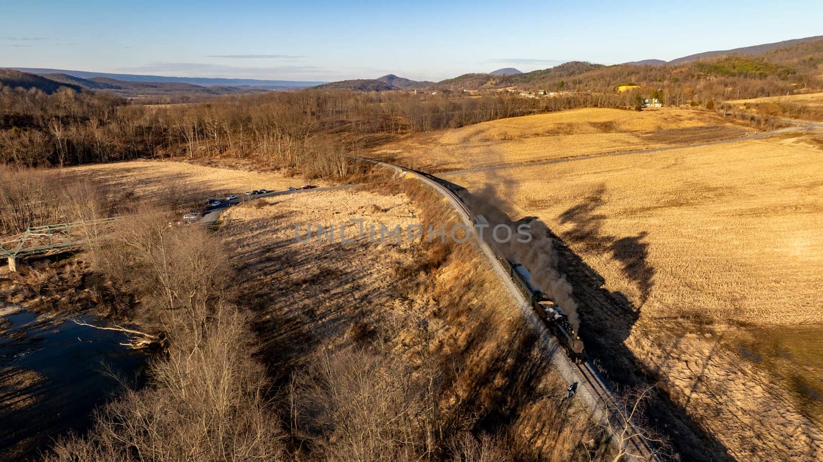 A vintage train winds along a curved railway surrounded by open fields and trees under a clear sky. The train creates a plume of smoke as it makes its journey through the peaceful countryside.