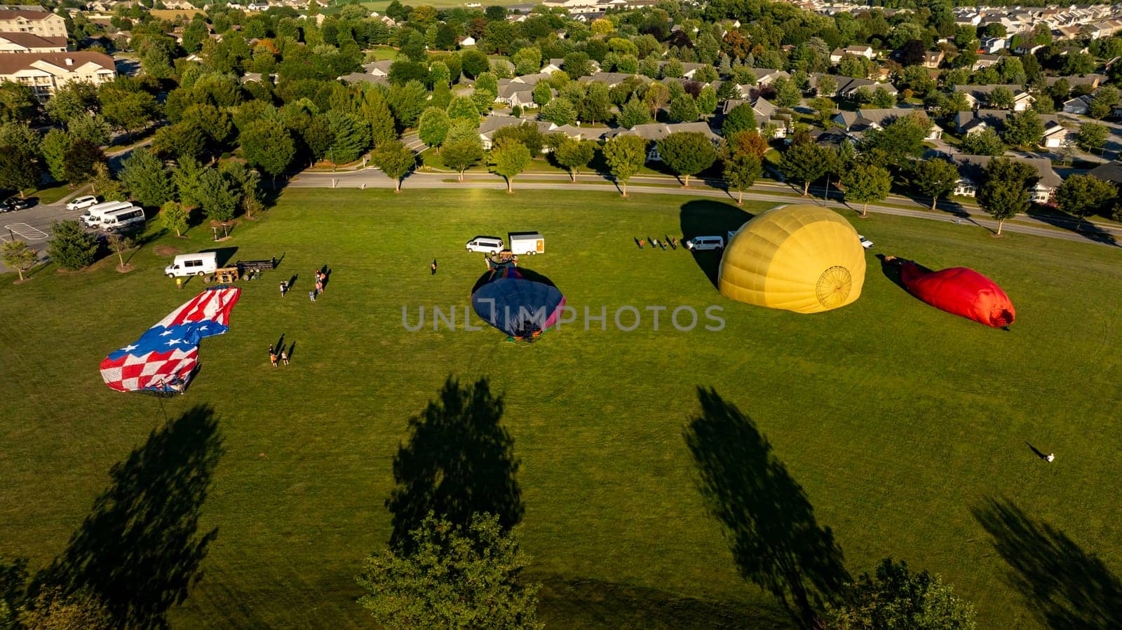 Brightly colored hot air balloons are laid out on the grass in a suburban park as teams prepare for an early morning ascent, surrounded by trees and houses.
