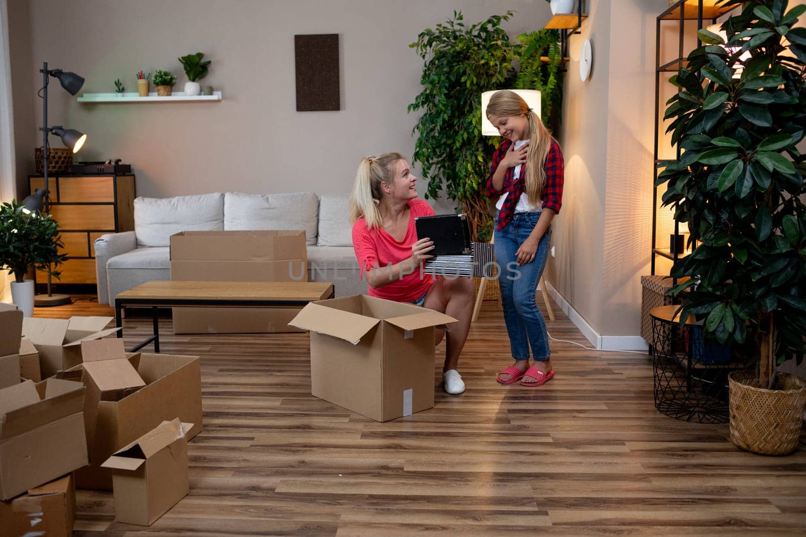 Two cheerful women are actively unpacking boxes in a bright, cozy living room that is filled with vibrant plants