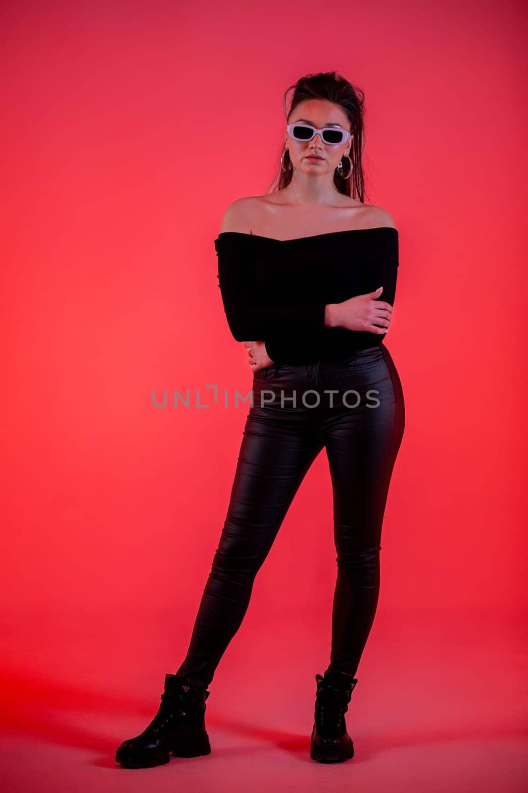 A confident woman proudly poses in an offshoulder top and stylish sunglasses against a vibrant red backdrop
