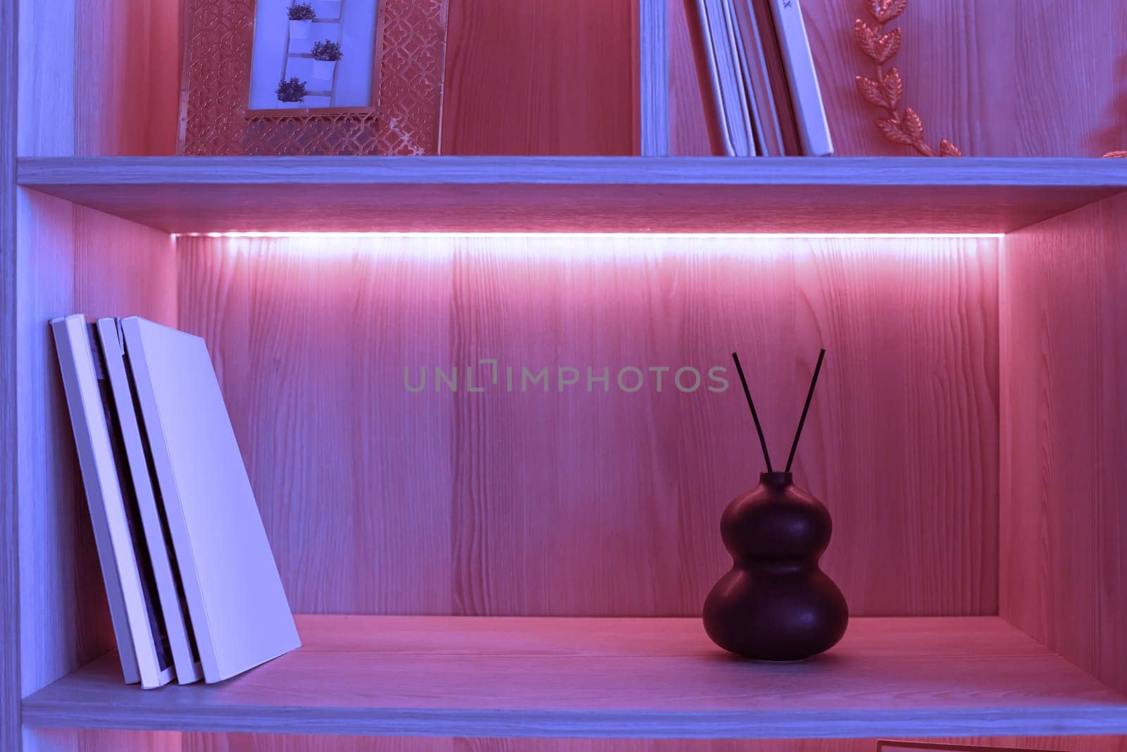 On a shelf with pink neon lighting there is a small black ceramic vase with two aroma sticks . Copy space