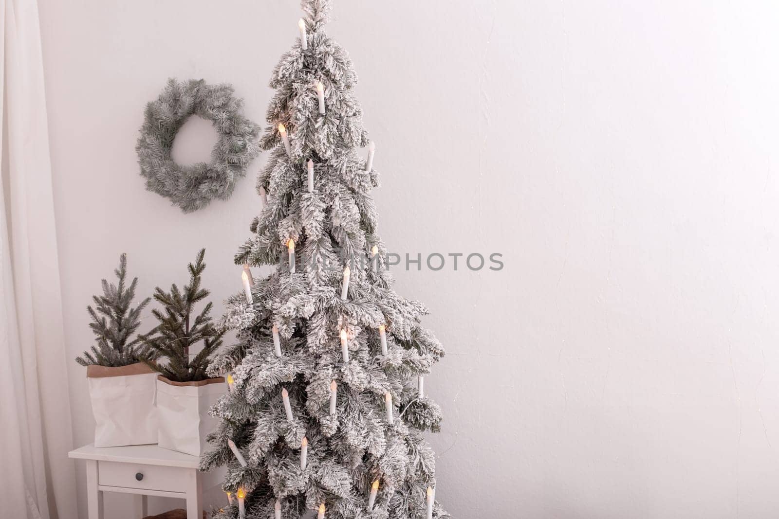 Snow-covered artificial spruce with an electric garland in the form of candles, standing against a white wall, at room. Copy space.