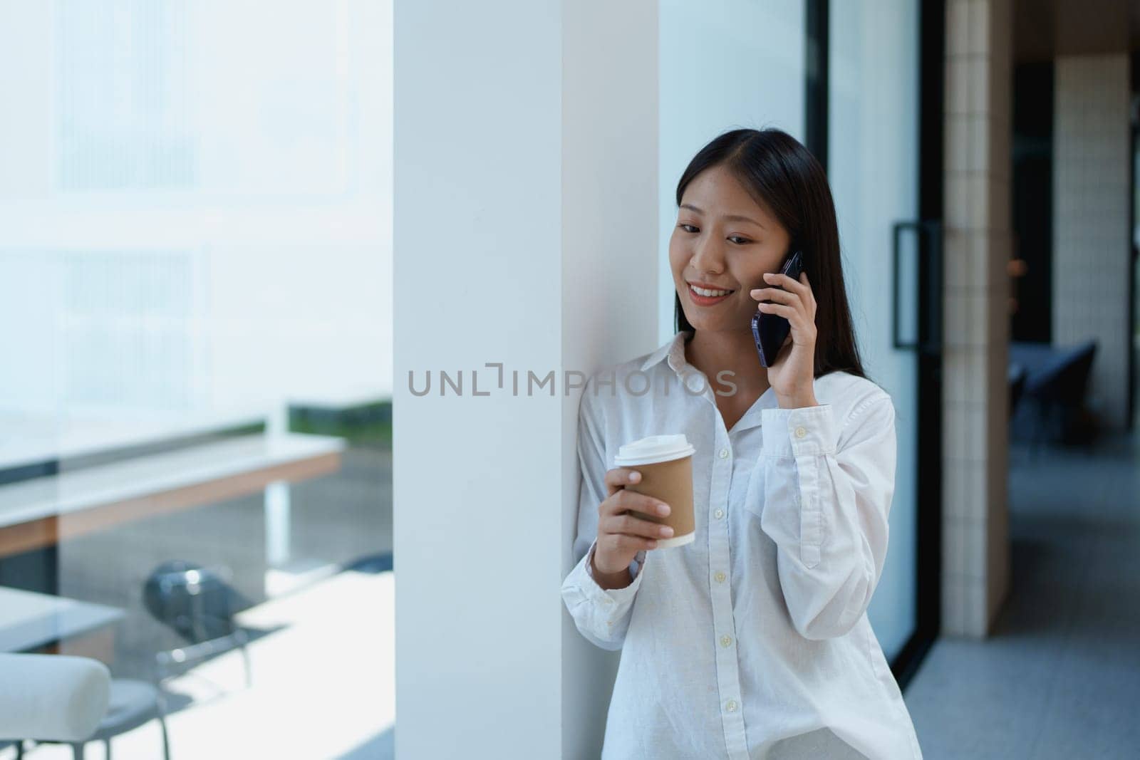 Young woman enjoying coffee while talking on a smartphone in a modern, bright office space with natural light.