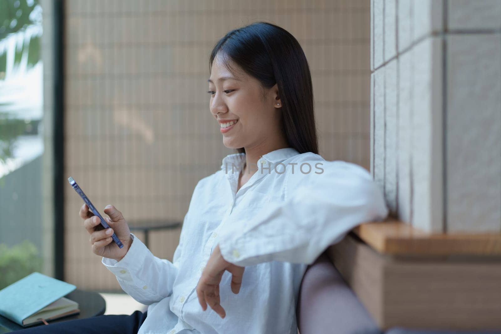Young woman sitting in modern cafe engaging with smartphone, smiling cheerfully amidst bright natural light.