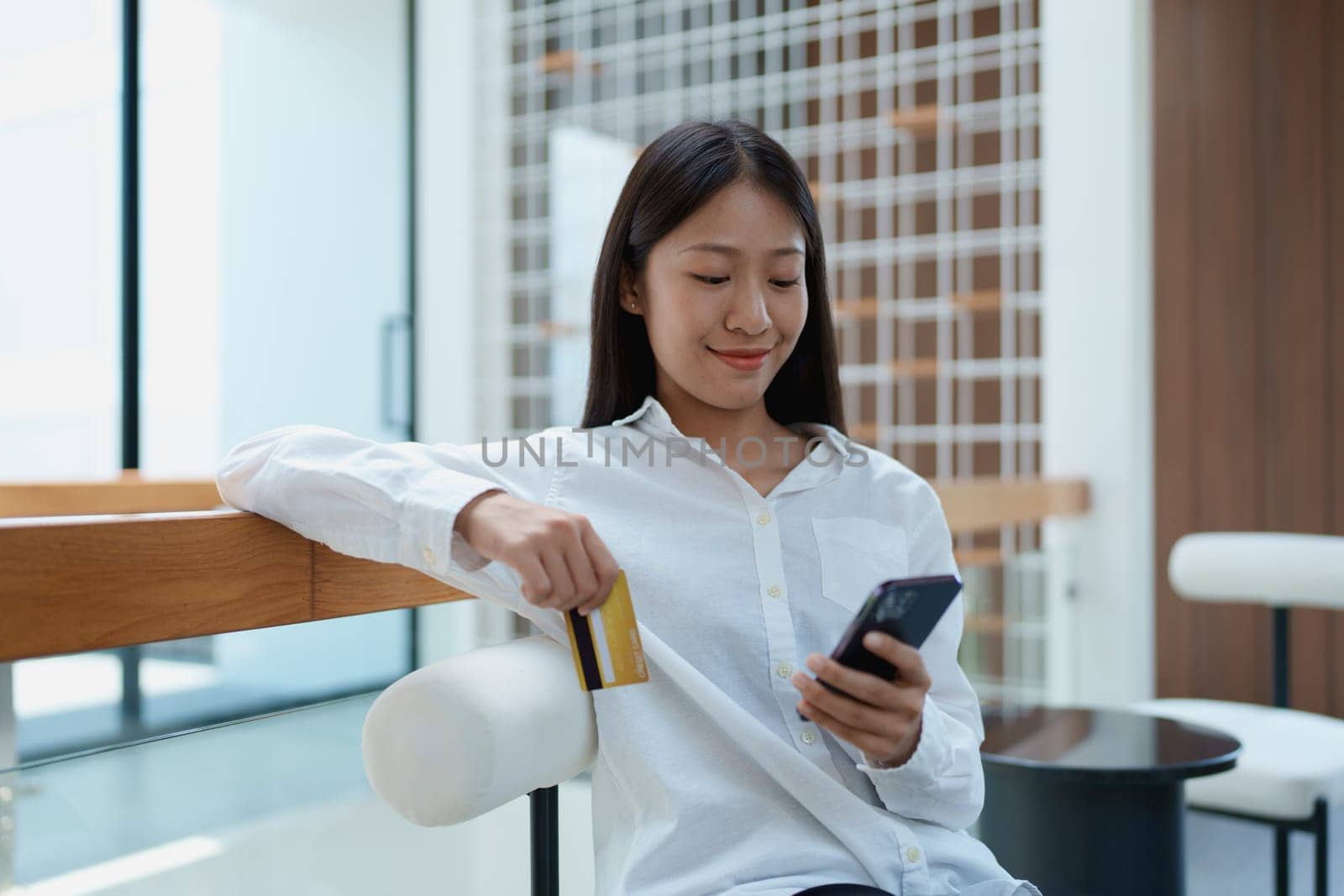 Young woman sitting comfortably in modern space, using smartphone while holding credit card with a smile.