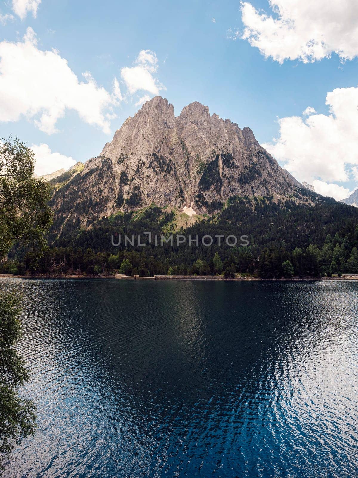 spectacular landscape of calm lake surrounded by massive green mountains and coniferous trees against blue sky in Aiguestortes i Estany of Saint Maurici National Park