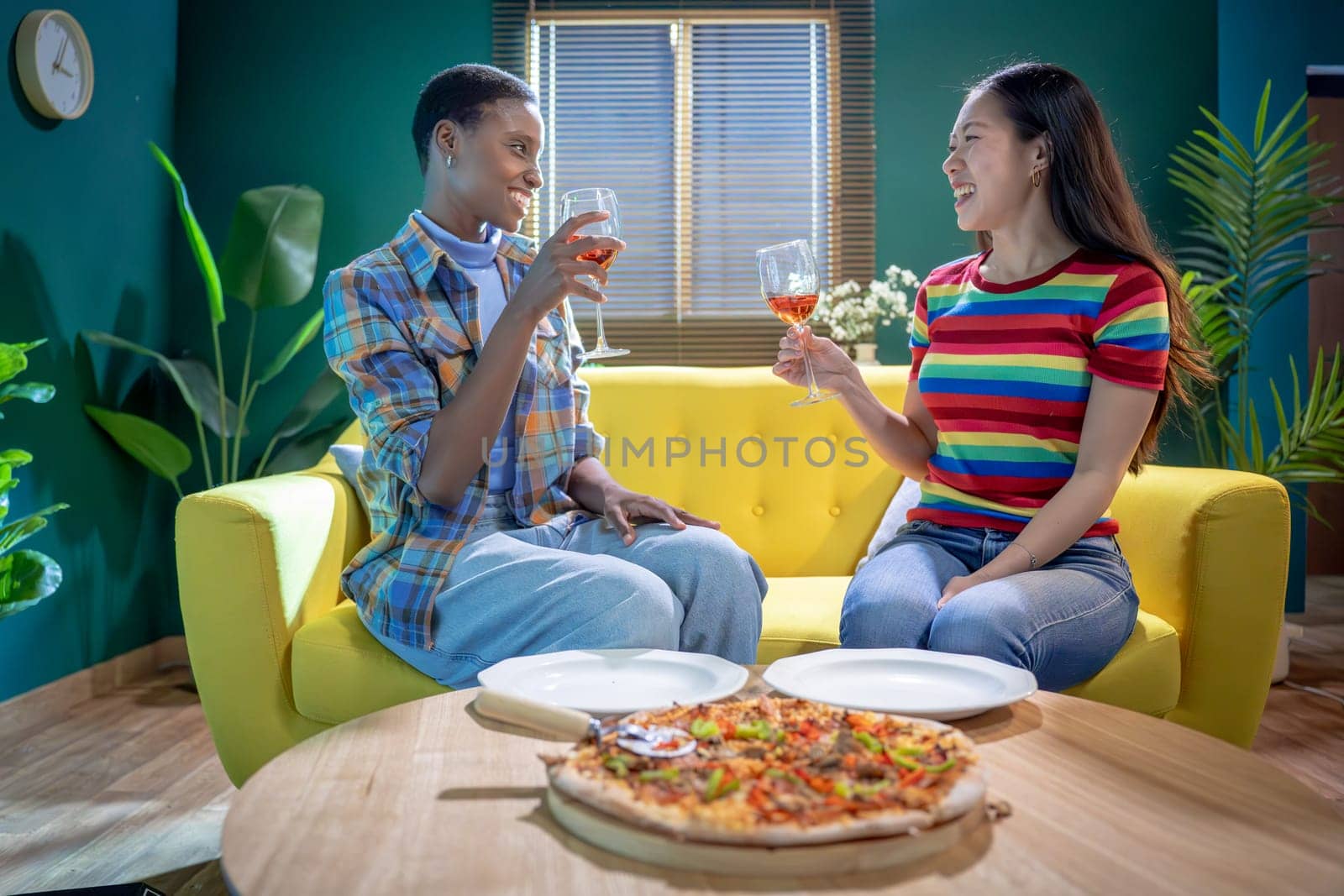 Two friends celebrate together with pizza and drinks on a cozy couch in a vibrant, colorful living room