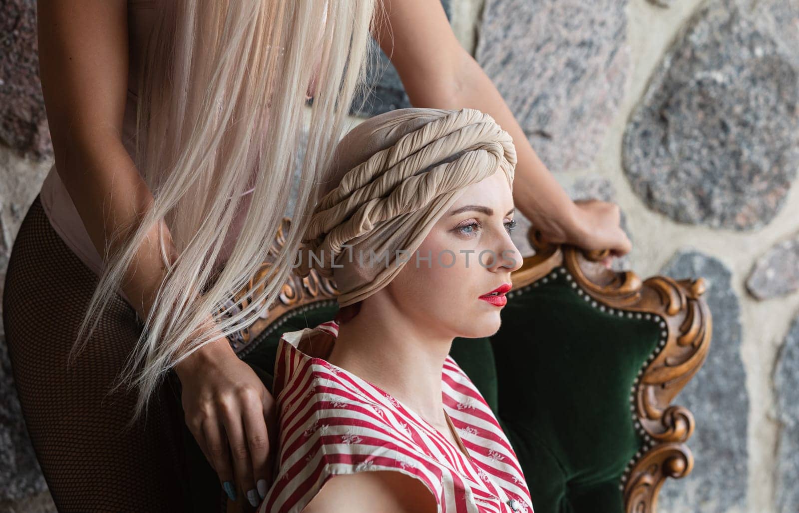 A stylist carefully prepares a unique hairstyle on a seated model. The rich textures of the stone wall complement the vintage chair, adding elegance to the creative process in soft lighting.