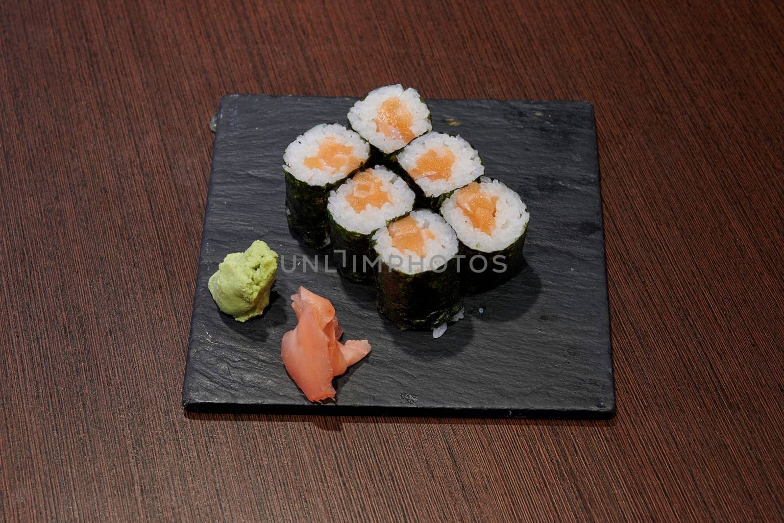 A plate of sushi with a green sauce and a piece of ginger on it. The sushi is rolled up and sitting on a black plate