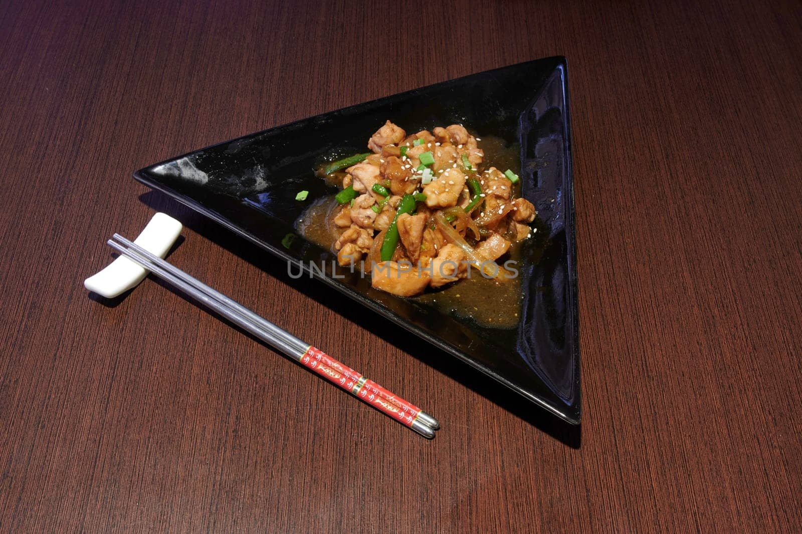 A plate of food with chopsticks on a wooden table. The plate is triangular and filled with chicken and vegetables