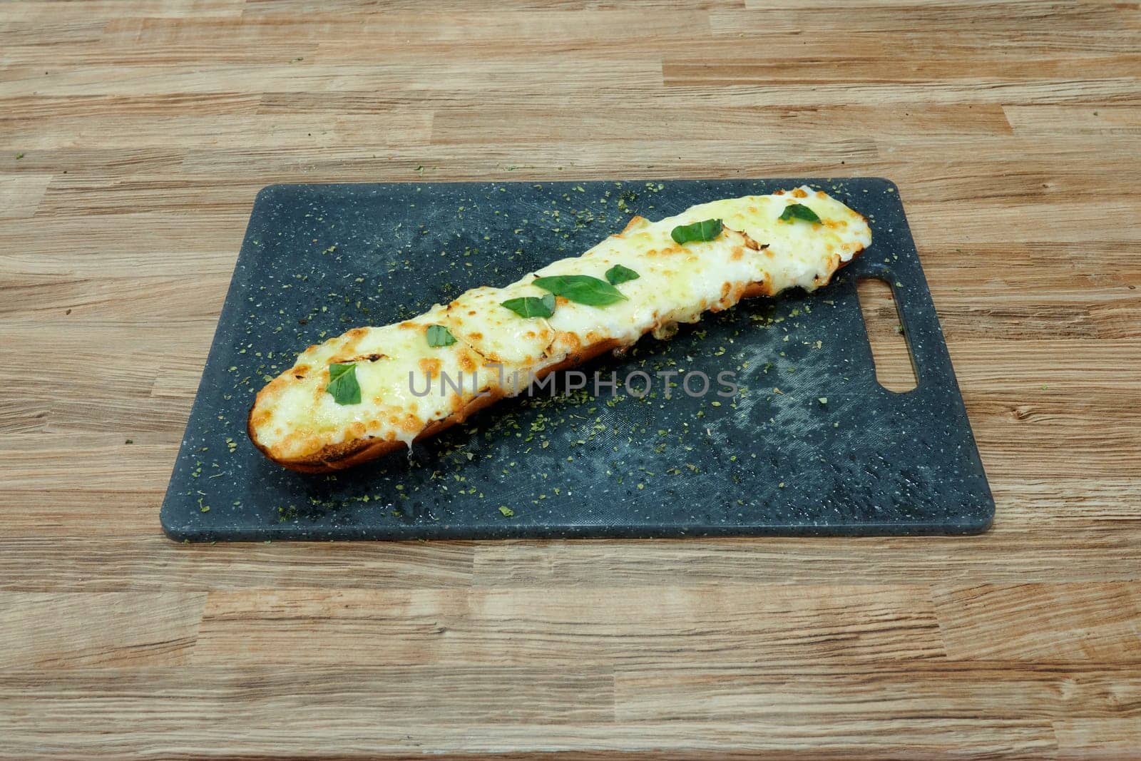 A long piece of bread with cheese and herbs on top. The bread is on a wooden cutting board