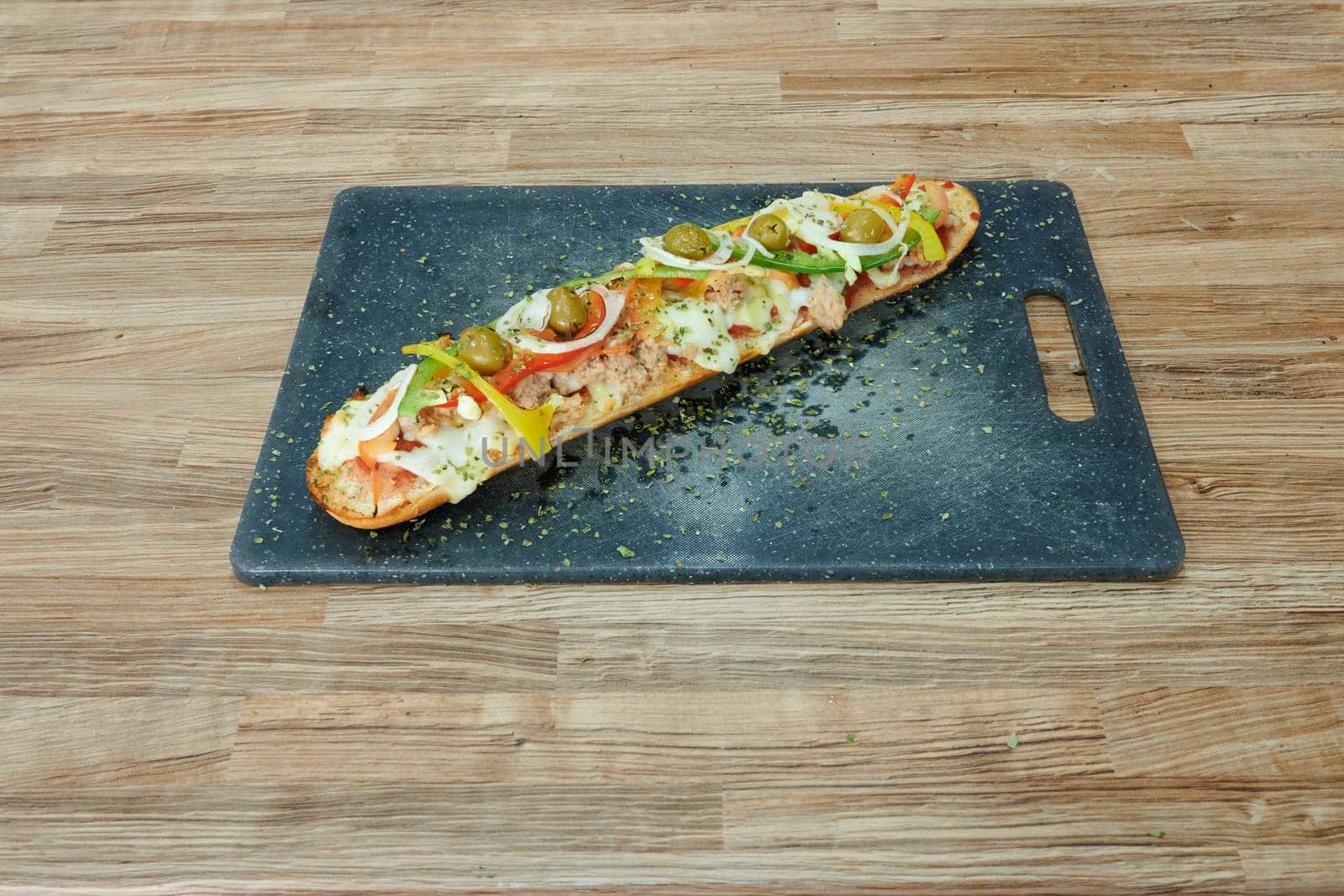 A long, thin pizza with a variety of toppings sits on a wooden cutting board. The pizza is topped with tomatoes, onions, and peppers, and it is a delicious and healthy meal