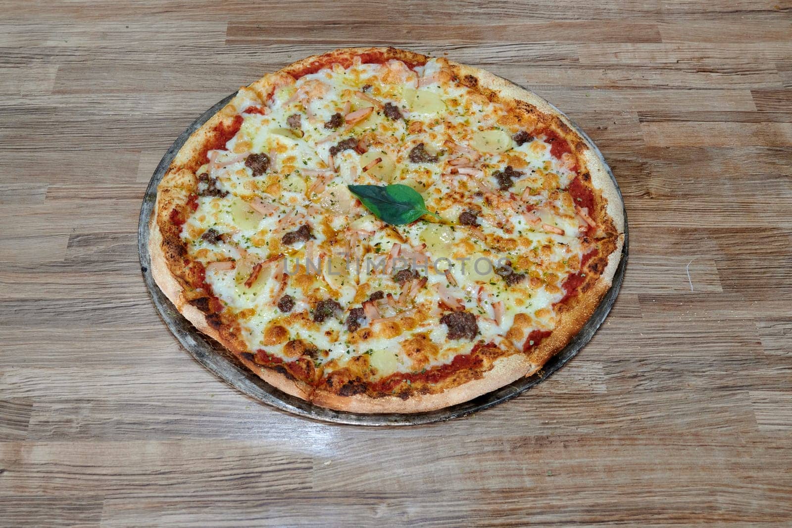 A pizza with a green leaf on top sits on a wooden table. The pizza is topped with cheese and meat, and it looks delicious