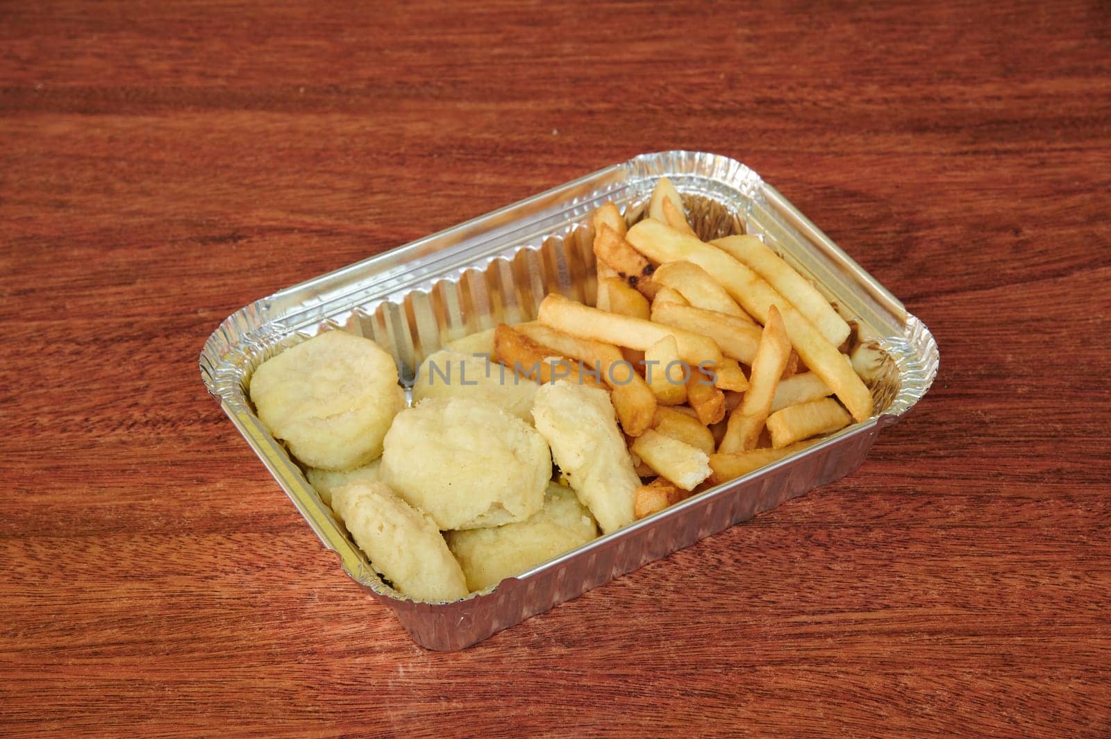 A tray of chicken nuggets and french fries sits on a wooden table. The tray is covered in aluminum foil, which is a common way to keep food fresh and prevent it from getting too greasy