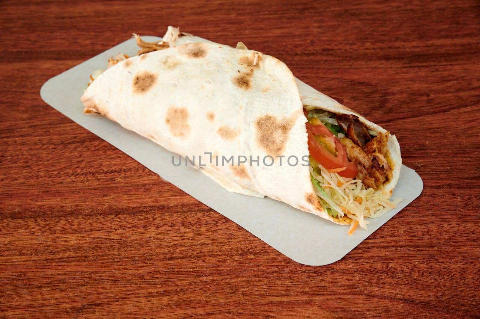 A wrap of food with lettuce and tomatoes on a wooden table. The wrap is on a white plate