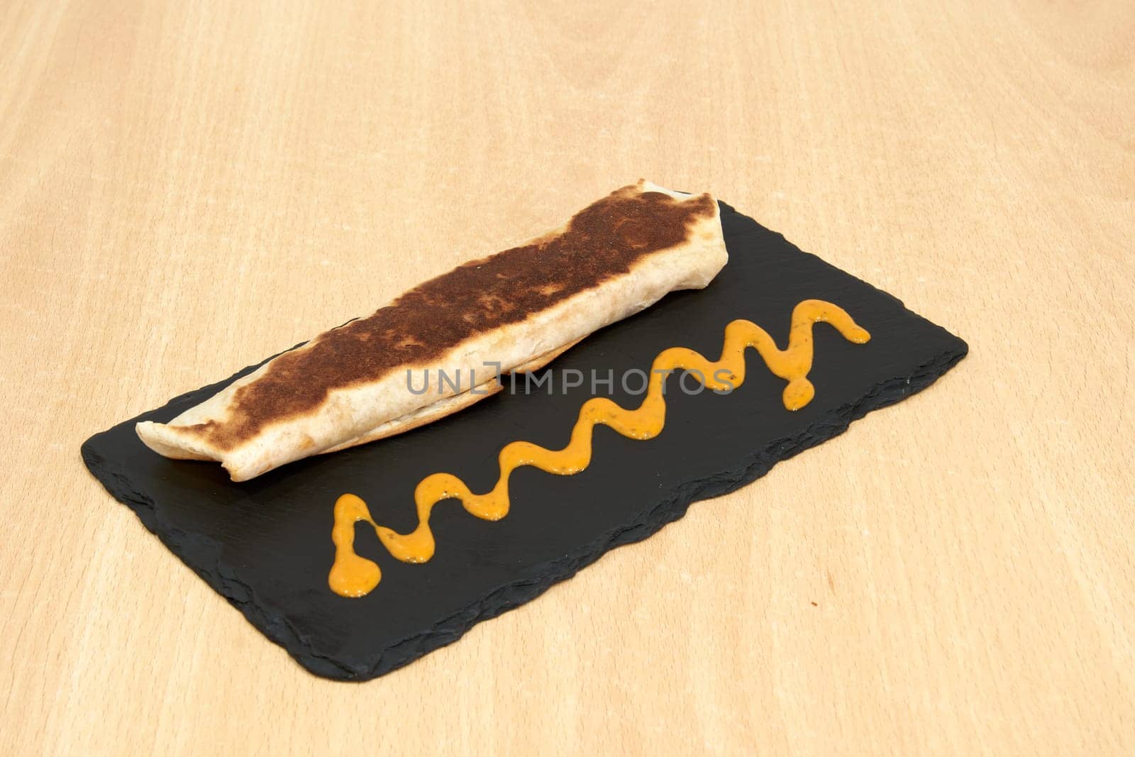 A tortilla wrap with a sauce on top of a black plate. The sauce is yellow and has a wavy appearance