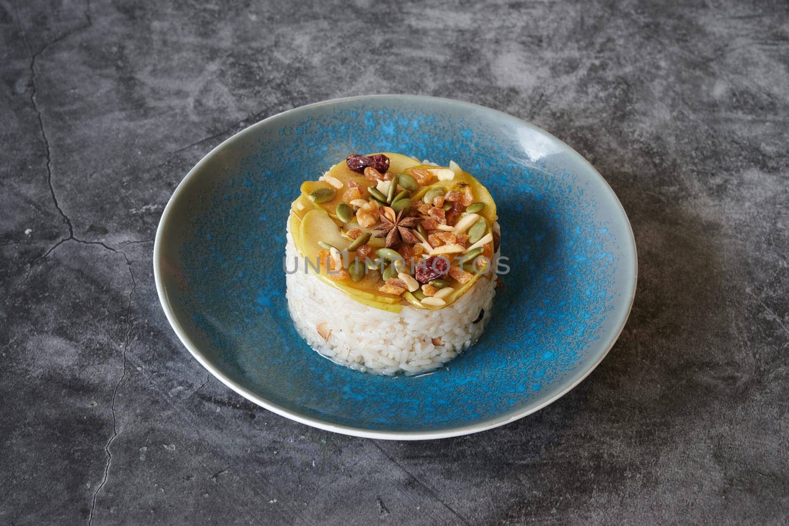 A blue plate with rice and nuts on it. The plate is on a grey surface. The rice is surrounded by nuts and there is a small piece of fruit on the plate
