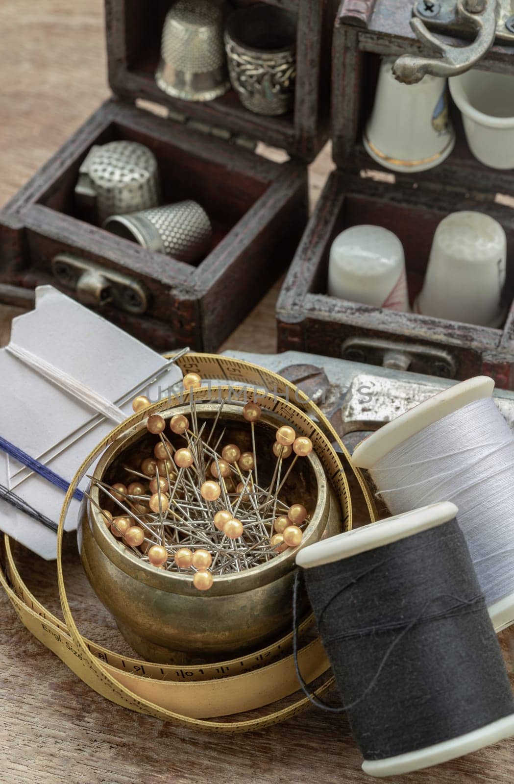 Sewing needles stuck in a white paper, Thimbles in small wooden treasure chest, Spools of black and white thread, Measuring tape and Steel scissors including pins in brass cup on wooden background. Sewing items set or Sewing objects (Sewing notions), use it as your Wallpaper, Poster and Space for text, Selective focus.