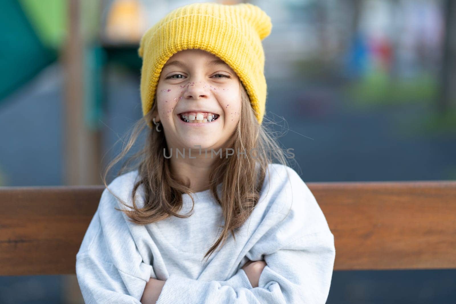 Portrait of cheerful pretty girl in yellow cap with freckles on face looking at camera while sitting on bench